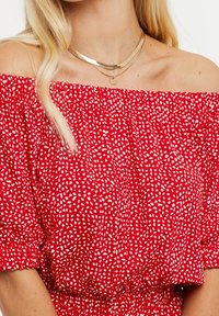 Haut rouge à épaules dénudées avec un design smocké, motifs abstraits blancs, poignets élastiques et une coupe confortable. Plusieurs colliers dorés superposés.