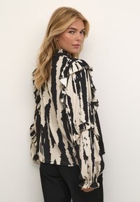 Blusa a righe nere e beige con maniche arricciate, collo alto e polsini arricciati; tessuto leggero con una texture liscia.