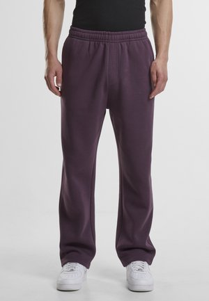 Muž v tmavofialových teplákoch a bielych teniskách stojí pred jednofarebným svetlým pozadím.