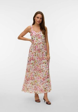 Femme debout, une main sur la hanche, portant une robe sans manches à imprimé floral, longueur cheville, et des sandales noires à talons.