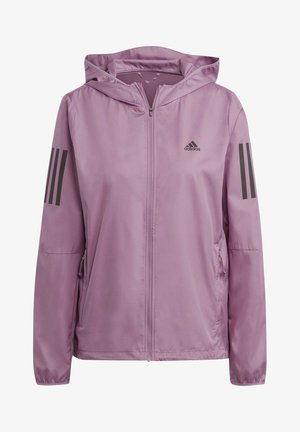 Kobieta ubrana w jasnofioletową bluzę Adidas na zamek z kapturem, z rękami w kieszeniach i dopasowane legginsy.