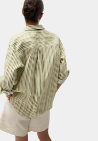 Camicia oversize a righe verdi e bianche con colletto, maniche arrotolate e schiena plissettata. Abbinata a pantaloni corti beige chiaro.