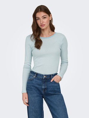 Jeune femme aux longs cheveux bruns, portant des boucles d'oreilles créoles, une chemise bleu clair texturée à manches longues et un jean bleu taille haute, la main dans la poche.