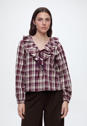 Vrouw draagt een bordeauxrood en wit geruit blouse met geplooide kraag en strik, gecombineerd met donkere broek, staand tegen een effen achtergrond.
