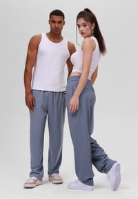 Deux individus portant des débardeurs blancs ajustés et des jogging gris à côtes. Leur chaussure comprend des baskets blanches et des chaussures de couleur claire.