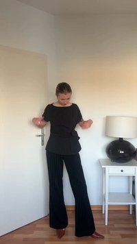 Γυναίκα διορθώνει μαύρο μπλούζα με ρίγες και παντελόνι, στέκεται κοντά σε λευκή πόρτα και λευκό πλευρικό τραπέζι με μαύρο φωτιστικό σε φωτεινό δωμάτιο.