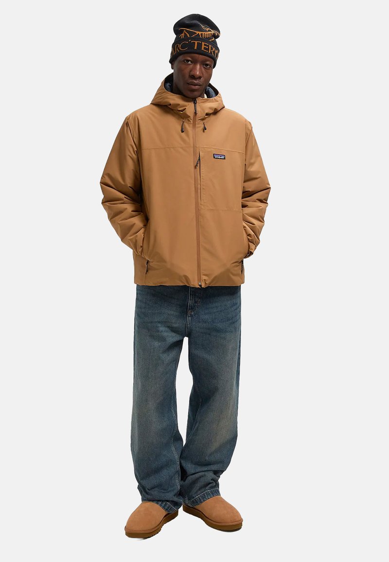 Giacca isolante marrone con cappuccio, jeans in denim blu e scarpe slip-on color cammello. Berretto nero con logo arancione.
