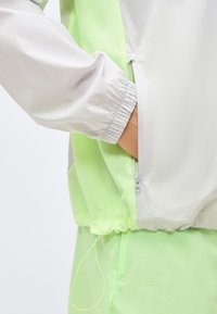 Χέρι με ανοιχτόχρωμο δέρμα που ακουμπάει μέσα σε φερμουάρ τσέπης ελαφρώς πράσινου και λευκού μπουφάν με ελαστική μανσέτα και κορδόνι στη μέση.