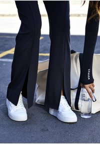 Čierne športové nohavice so štrbinami na bokoch, doplnené bielymi teniskami. Osoba drží priehľadnú fľašu s vodou vedľa béžovej tašky.