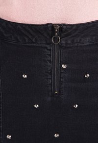 Minifalda de mezclilla negra con cremallera frontal, adornada con tachuelas plateadas distribuidas uniformemente. La tela tiene una textura suave.