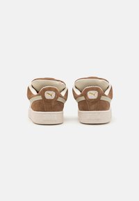 Brown suede sneakers with white accents, textured rubber sole, and a gold logo on the heel. Minimalist design with padded collar.