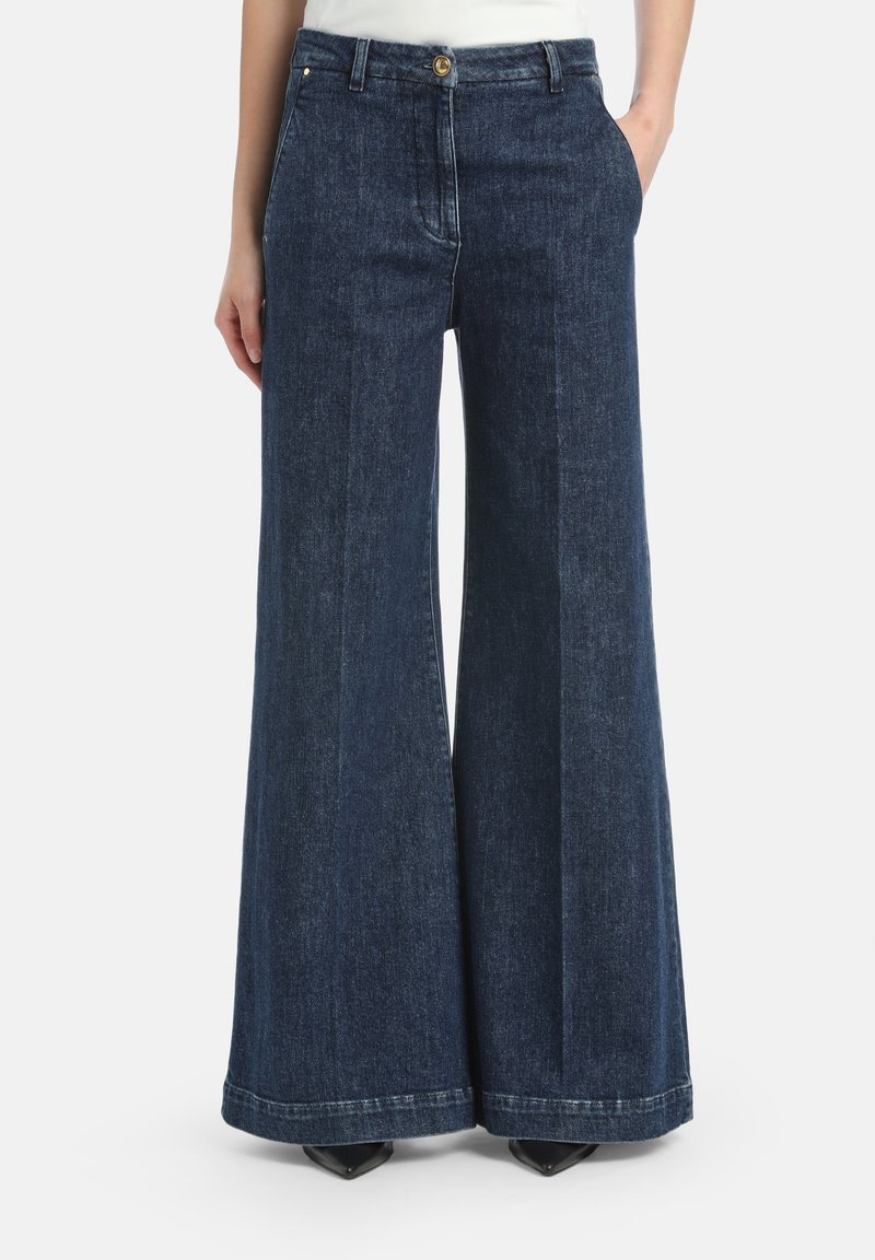 Vrouw in een donkerblauwe, hoge taille, wijde denim jeans met zakken en een zichtbare knoopsluiting.