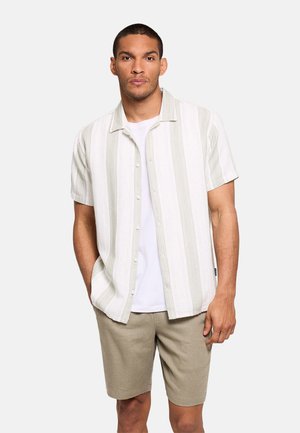 Hombre con camisa de manga corta a rayas color beige claro sobre una camiseta blanca, con pantalones cortos beige, de pie frente a un fondo liso.