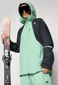 Mintgrønn og svart ski-jakke med hette, glidelåslommer og branding. Ski og bindinger holdt i venstre hånd, svarte goggles brukt.