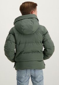 Groene gewatteerde jas met een capuchon, geribbeld kraag, zijzakken en een logo-detail op de bovenrug. Draagt over lichtblauwe jeans.