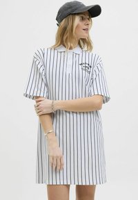 Robe polo rayée blanche avec des rayures verticales noires, manches courtes et une patte de boutonnage. Présente un logo sur la poitrine.