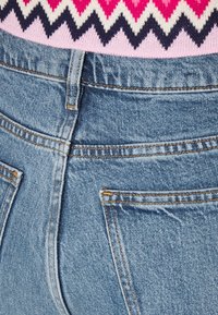 Ljusblå denimjeans med hög midja, förstärkta sömmar och två synliga bakfickor, mot en topp i rosa med zick-zack-mönster.