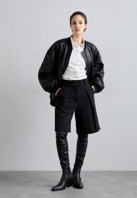 Black leather bomber jacket over a white shirt, paired with high-waisted black shorts and thigh-high black leather boots. Smooth textures and tailored design.