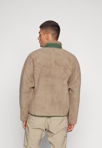 Fleece jacket in tan with a textured finish, high collar in green, and a relaxed fit. Back view shows stitching details and hemline.