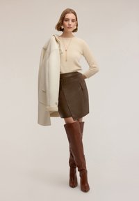 Beżowy wełniany sweter, brązowa skórzana spódnica z zakładką i boczną kieszenią oraz brązowe skórzane kozaki za kolano na obcasie, zestawione z kremowym płaszczem.