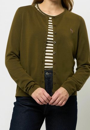 Femme portant un cardigan vert olive boutonné par-dessus une chemise blanche à rayures olive et un jean bleu foncé, mains jointes à la taille.
