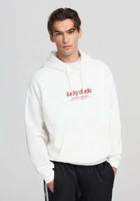 Felpa bianca con cappuccio, tasca frontale e stringhe. Presenta un logo rosso stampato con la scritta "lucky dude" e un testo sottostante. Design semplice e texture morbida.