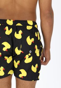 Zwarte zwembroek met een herhaald patroon van gele rubberen eendjes met rode accenten. Inclusief een achterzak met een drukknoopdetail.