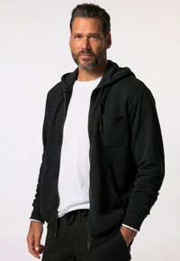 Sudadera negra con cremallera, hecha de tela suave, que presenta un bolsillo frontal, capucha con cordón y puños acanalados. Lleva puesta una camiseta blanca.