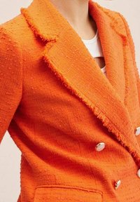 Oranžový kostkovaný dvouřadý blejzr s roztrhanými klopami a bílými knoflíky, nošený přes bílý top.