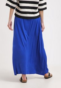 Longue jupe maxi bleue en tissu lisse, avec une taille froncée et un design fluide. Associée à des sandales noires pour un look décontracté.