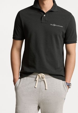 Schwarzes Polohemd mit strukturiertem Gewebe, ausgestattet mit einer dreiknöpfigen Knopfleiste und einem Logo. Kombiniert mit hellgrauen Jogginghosen und einer Uhr.