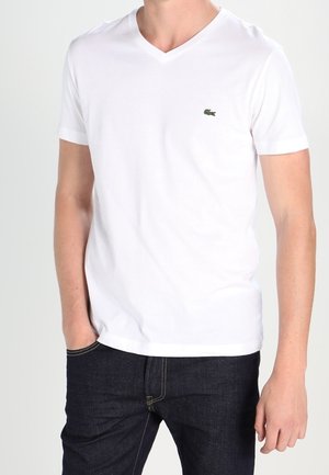 T-shirt bianca in cotone con scollo a V, maniche corte e un piccolo logo verde Lacoste sul petto; abbinata a jeans in denim scuri.