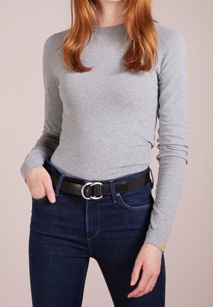 Personne portant une chemise grise à manches longues rentrée dans un jean bleu foncé avec une ceinture noire, debout avec une main dans la poche.
