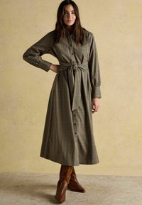 Garšs pogājušais kleita ar zaļu un brūnu rūtainu rakstu. Īpašības ietver sasietu vidukli, garas piedurknes un to papildina brūnas papēžu zābaki.