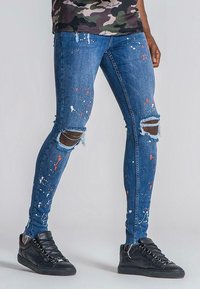 Smala denimjeans i medelblått med färgstänk, med slitna knäskärningar och fransad avslutning. I kombination med svarta sneakers.