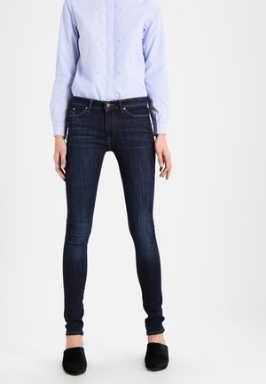 Personne portant un jean skinny bleu foncé associé à une chemise à manches longues rayée bleu clair rentrée à l'intérieur et des chaussures noires sans lacets.