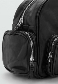 Mochila de cuero negro con cremalleras metálicas. Cuenta con múltiples bolsillos con cremallera, textura suave y forma curva. Diseño compacto.