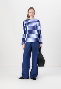 Blue and white striped long-sleeve shirt, paired with wide blue trousers, and black handbag. Model stands against a white background.