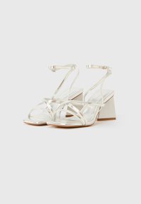 Silberne metallische Sandalen mit hohen Absätzen, Knöchelriemen, eckigen Absätzen und gekreuzten Riemen im Zehenbereich. Glatte, reflektierende Oberfläche.