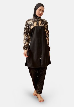 Vrouw draagt een zwarte tuniek met bloemenpatroon en lange mouwen, bijpassende losse zwarte broek en een zwarte hijab, staand op blote voeten tegen een effen achtergrond.