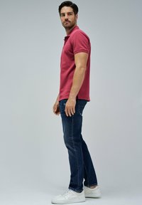 Hombre de perfil lateral vestido con una camiseta polo roja, jeans azul oscuro y zapatillas blancas, contra un fondo gris liso.