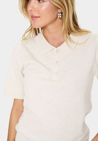 Beige poloko s krátkým rukávem a límečkem, se třemi knoflíky, vyrobené z texturované tkaniny, částečně viditelné na neutrálním pozadí.