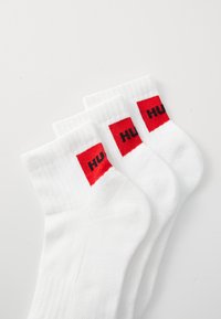 Tres pares de calcetines blancos con puños acanalados, que presentan acentos en rojo brillante y letras negras "HU" en cada calcetín. Textura suave y longitud media.