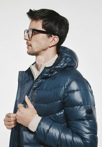 Granatowa kurtka puchowa z kapturem, o pikowanej fakturze i zamkiem błyskawicznym. Posiada kieszeń na piersi oraz elastyczne mankiety, noszona na beżowej koszuli.