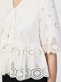 Blusa bianca in cotone con scollo a V, chiusura con laccetto e maniche a palloncino. Presenta un ricamo ad occhiello dettagliato e un orlo scallop.