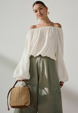 Femeie purtând o bluză albă cu umerii goi și mâneci bufante, pantaloni largi de culoare verde măsliniu și ținând o geantă împletită cu curele de piele.