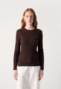 Pull en tricot marron avec un motif en relief, col rond et côtes aux poignets et à l'ourlet. Présente un petit logo sur la poitrine gauche.