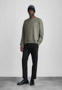 Olive green sweatshirt with ribbed cuffs, featuring a white graphic. Paired with black trousers and a knitted navy beanie. Slim fit shoes.
