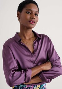 Camicetta di raso viola con colletto a bottoni, maniche lunghe arrotolate ai polsini, indossata con collane d'oro a strati e una gonna con fantasia.