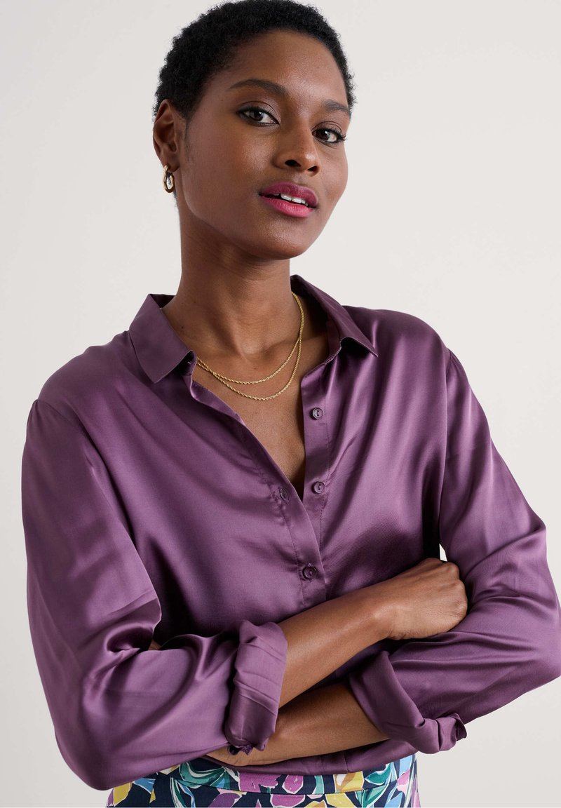 Camicetta di raso viola con colletto a bottoni, maniche lunghe arrotolate ai polsini, indossata con collane d'oro a strati e una gonna con fantasia.